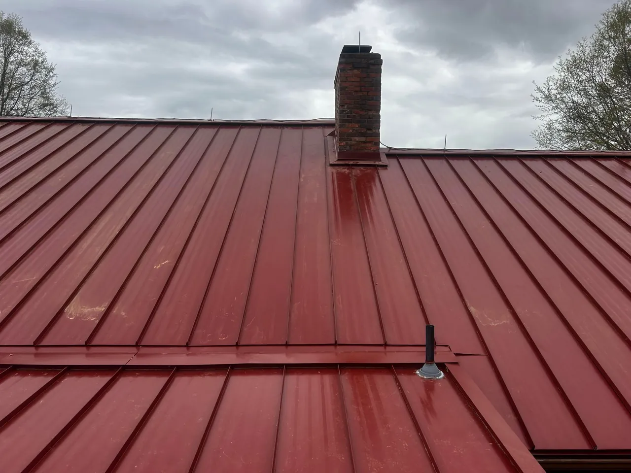 New red standing-seam metal roof with a brick chimney on a home in Benson, NC, under a gray sky