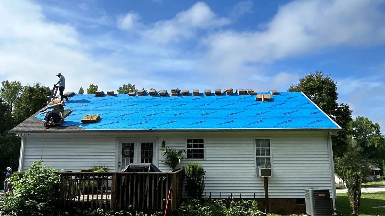 Crew installing new shingles over blue synthetic underlayment on a one-story home in Clayton, NC, with bundles staged along the ridge