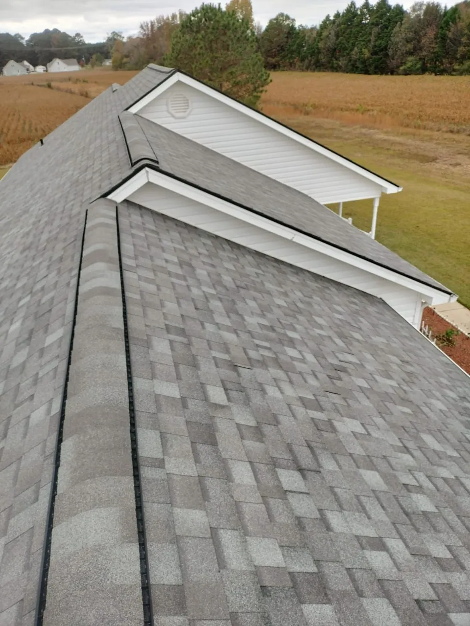 New gray architectural shingles with ridge vents on two ridges of a ranch home in Pine Level, NC, fields beyond
