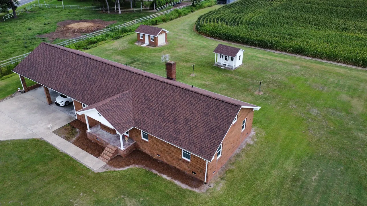 Drone view of a brick ranch home in Edenton, NC with a new brown architectural shingle roof, a carport and fields beyond
