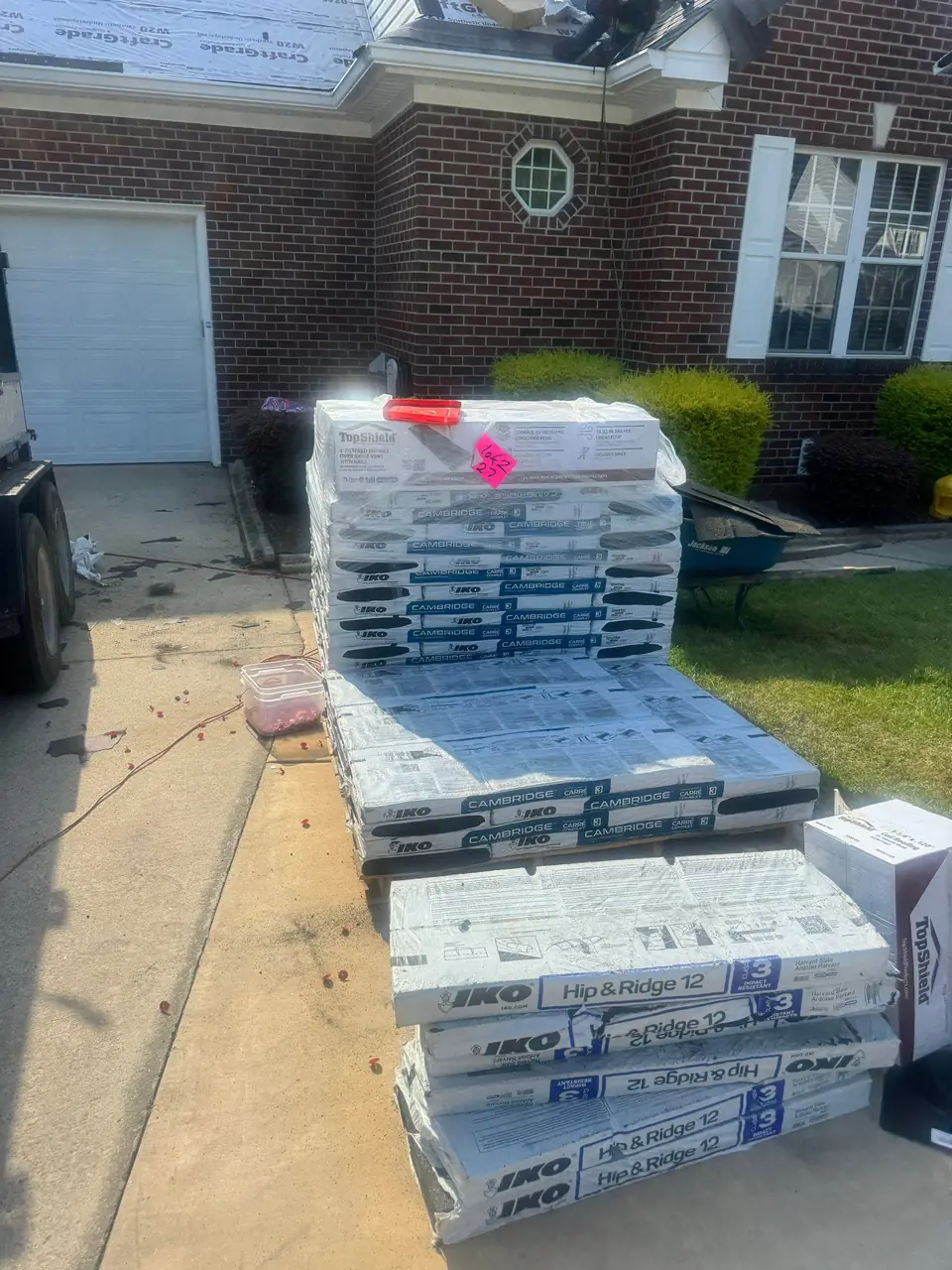 Pallets of IKO Cambridge shingles and IKO Hip & Ridge 12 cap shingles in a driveway in Goldsboro, NC, with the tear-off under way on the roof