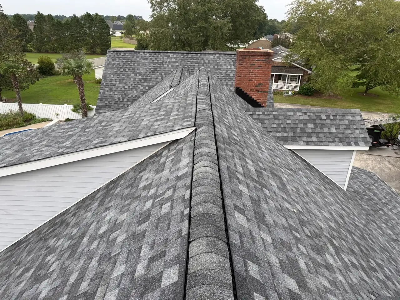 Finished gray architectural shingle roof in Greenville, NC with ridge caps along the hips and black step flashing at a brick chimney