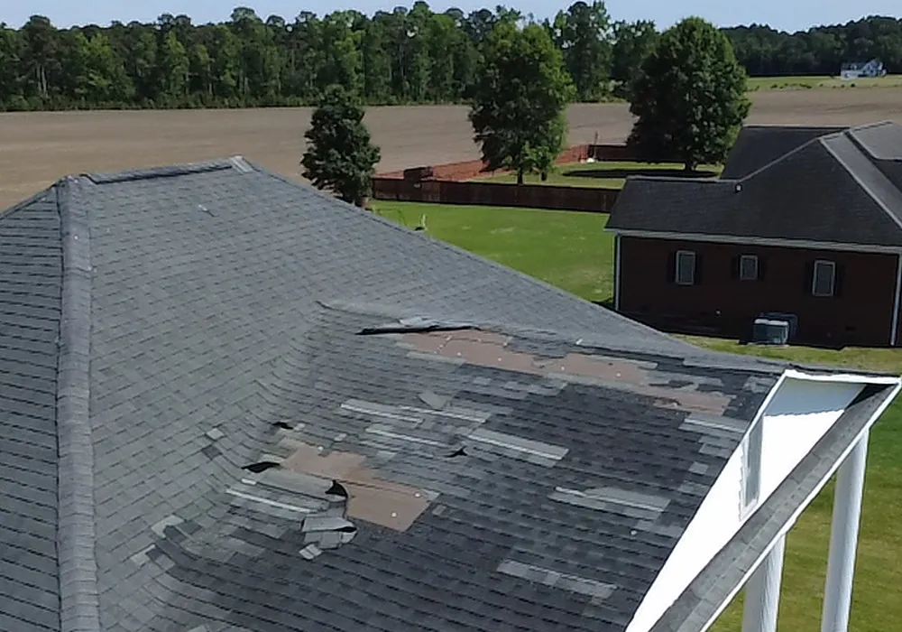 Aerial view of a shingle hip roof in Grimesland, NC with missing and displaced shingles above a white-columned porch, farm fields beyond