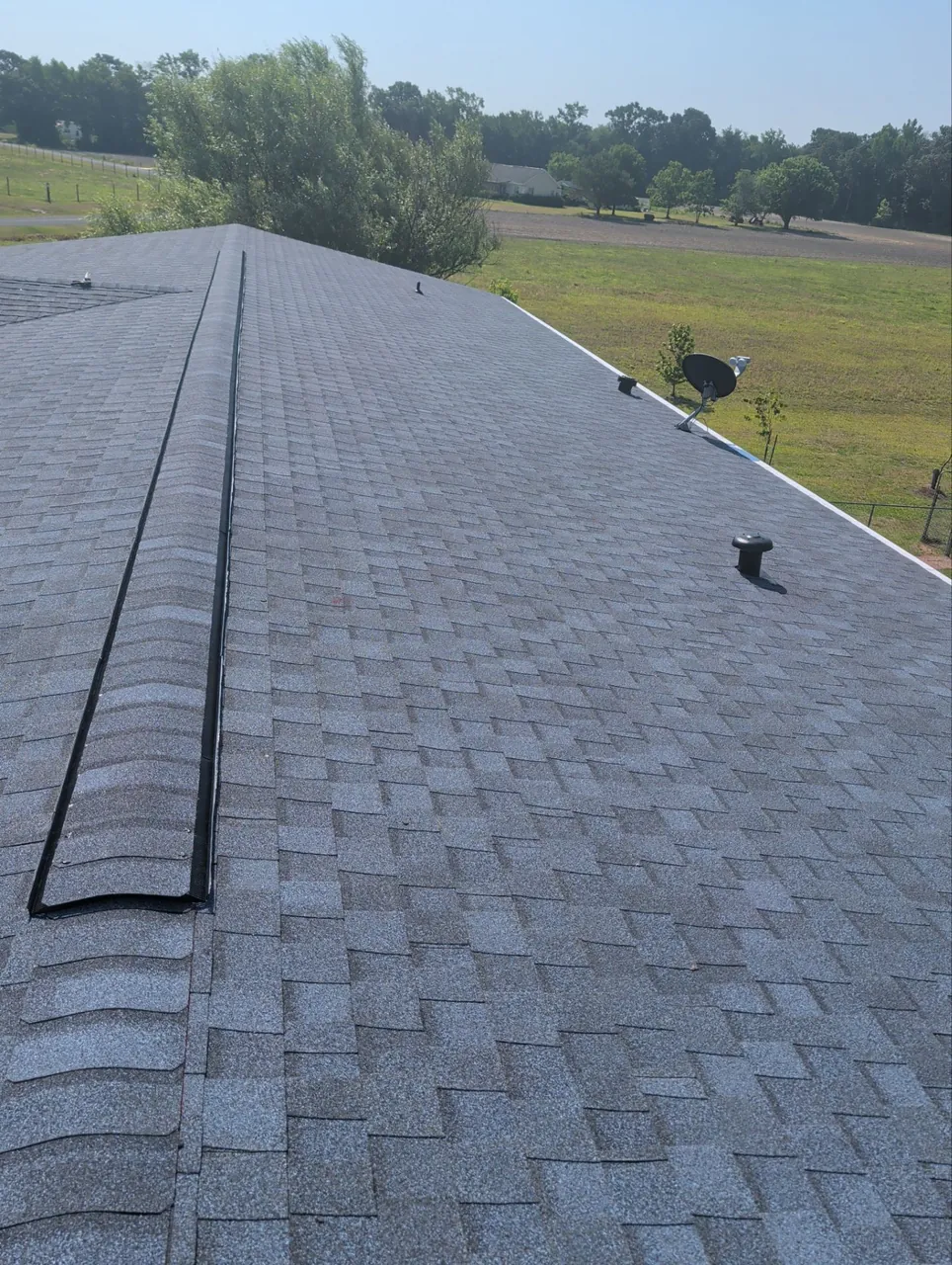 New gray architectural shingles, ridge vent and cap along the peak of a roof in Beulaville, NC, with open fields beyond