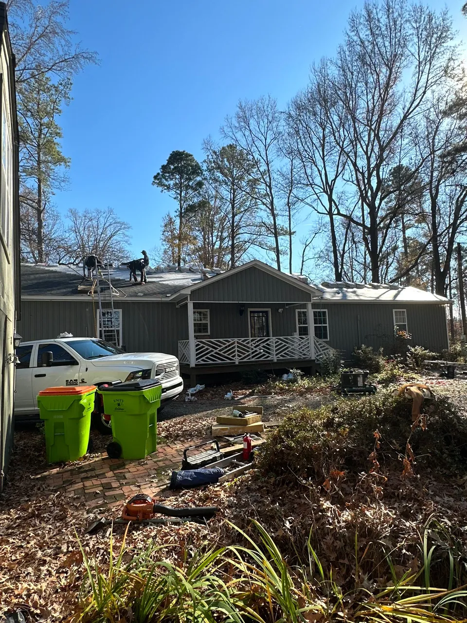 Roofers on top of a green house in Enfield, NC during the tear-off, with trash bins and tools in the yard