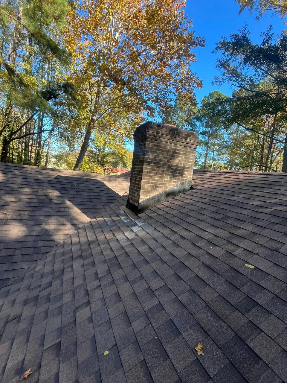 Brick chimney rising from a new brown architectural shingle roof in Roanoke Rapids, NC, autumn trees behind