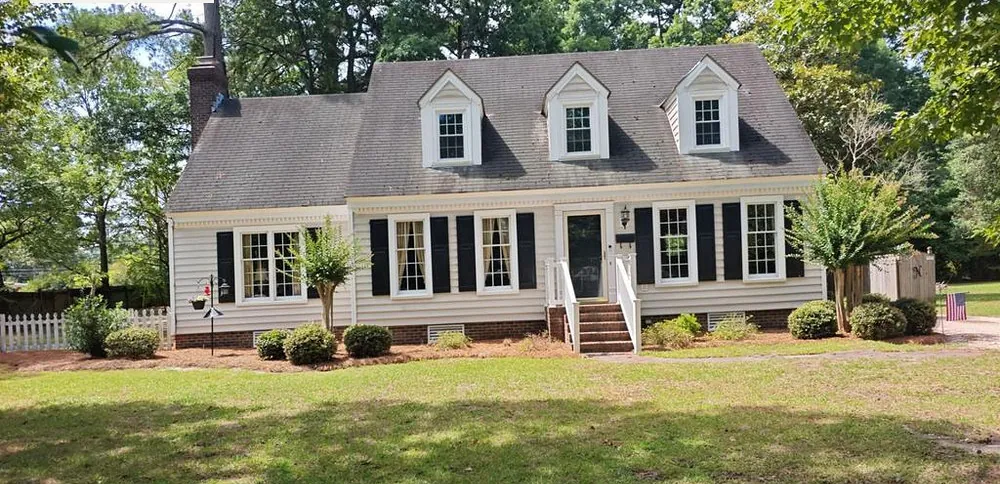 White Cape Cod home in Rocky Mount, NC with three dormers under an old, dark, streaked shingle roof, June 2026