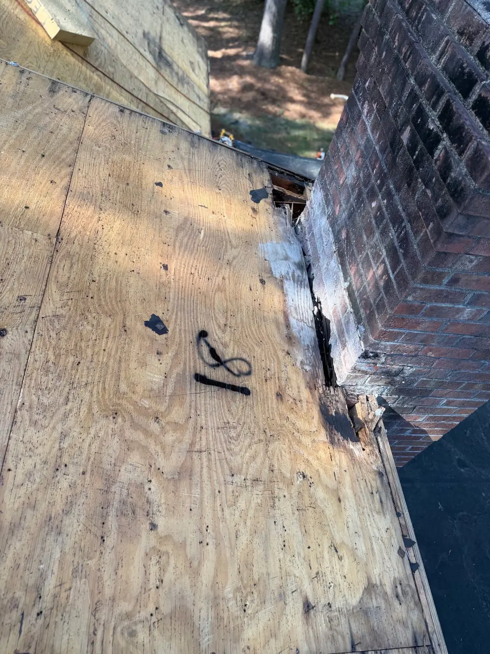 Bare plywood roof deck beside a brick chimney during a roof tear-off in Tarboro, NC, with the damaged deck edge along the chimney exposed