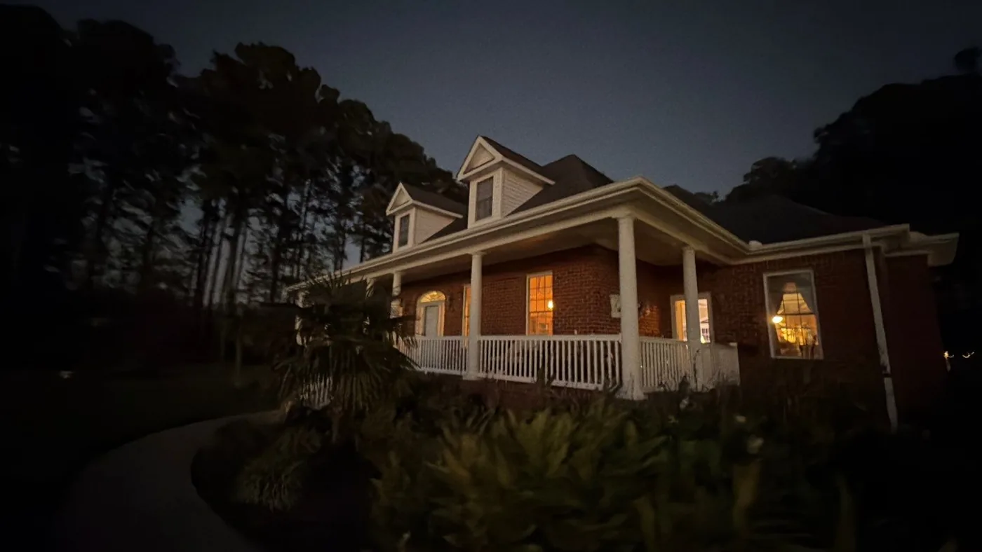 Two-story red brick colonial home with a white-columned wrap-around porch and dormer windows, photographed at dusk with the porch and interior lights glowing warm against a deep blue sky.