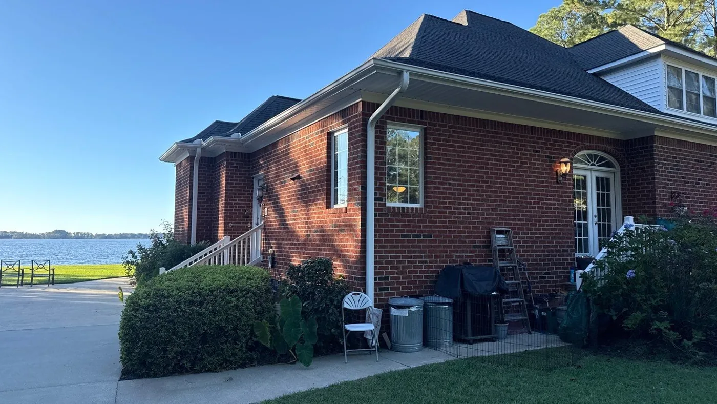 Waterfront elevation of a red brick home in full sun, showing the original dark asphalt shingle roof with hipped slopes, white gutters and downspouts, with open water and a grass lawn to the left.