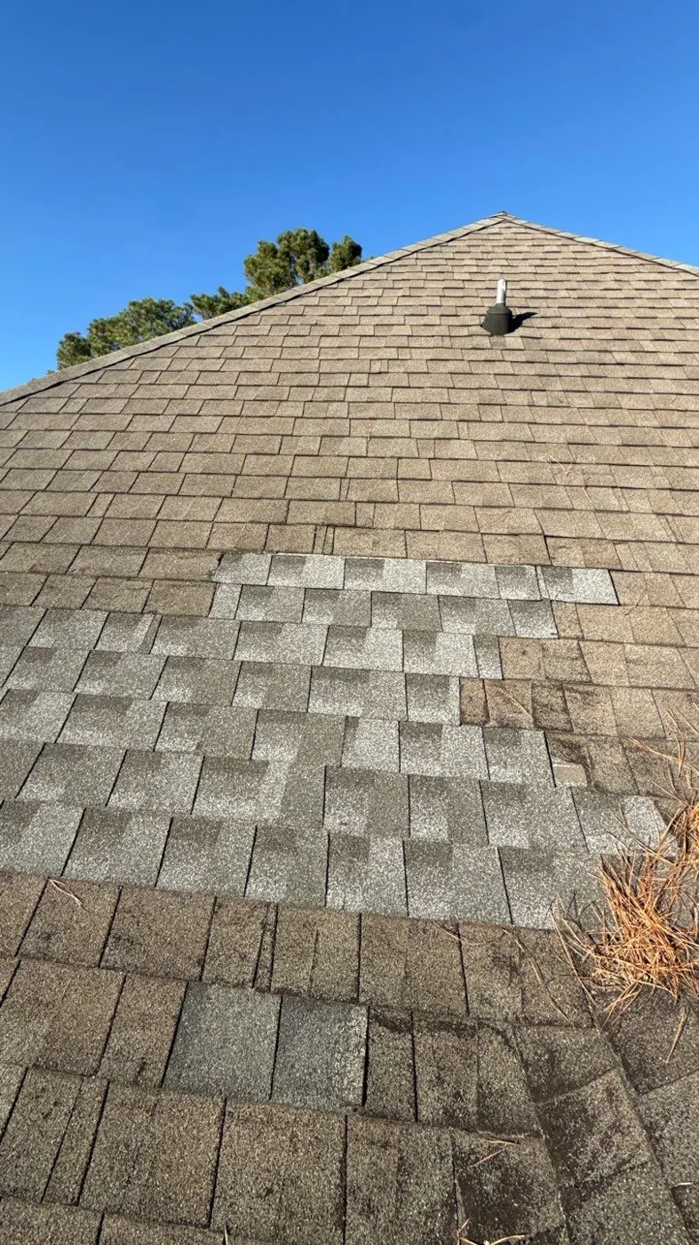 Close view up a sloped asphalt shingle roof, showing weathered grey-brown shingles with a large rectangular block of lighter, mismatched replacement shingles patched into the middle of the slope and pine needles collected in the courses.
