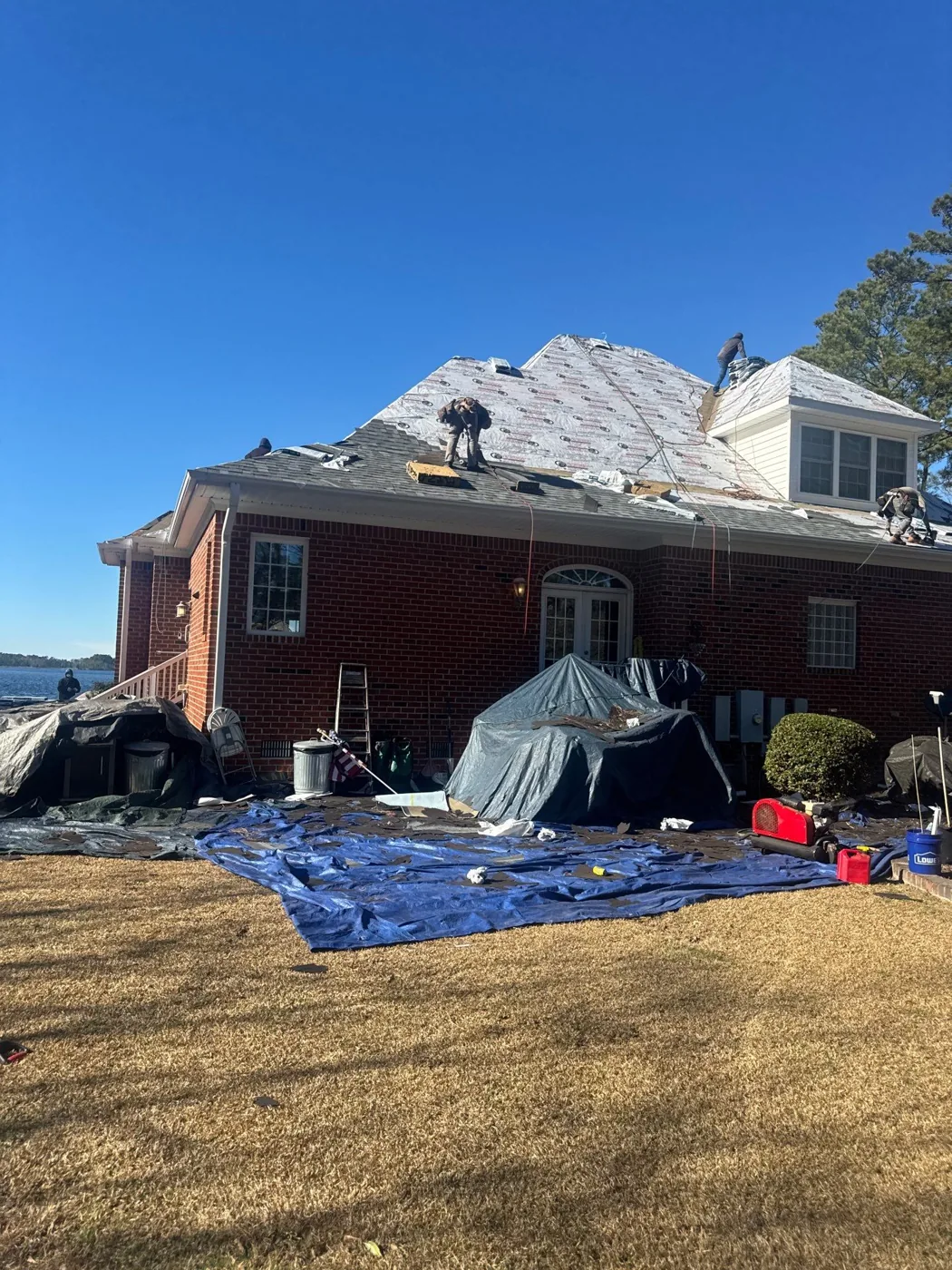 Side elevation of the red brick home with the roof partly stripped and covered in underlayment, crew members working on the slope above, grey tarps draped over shrubs and a large blue tarp spread across the lawn to catch debris, with water visible at the left.