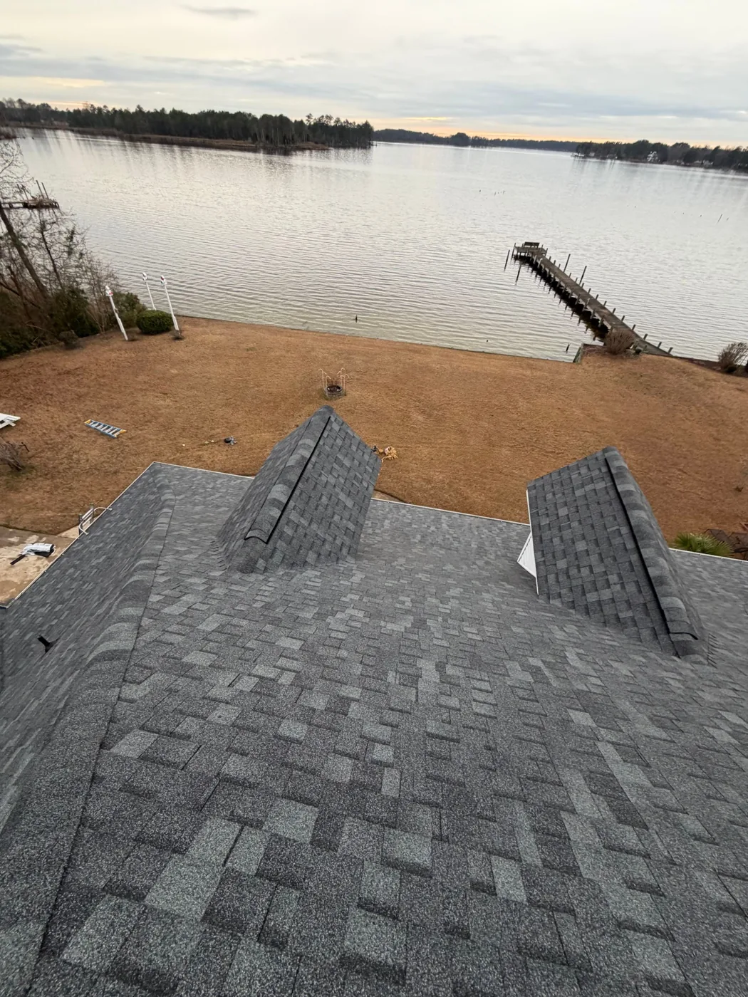 View from the ridge of the completed grey architectural shingle roof, looking over two shingled dormer peaks and hip ridges to a wide open bay with a long wooden pier and a treed shoreline in soft evening light.