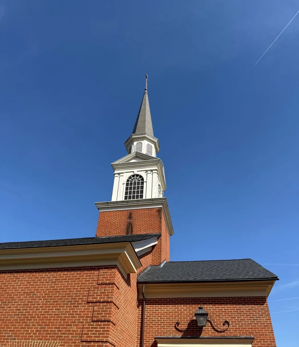 White multi-stage steeple with a tall spire rising above the brick tower of a church, seen from below against a deep blue sky.