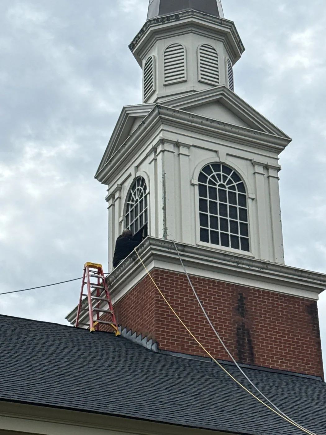 A step ladder set on a shingle roof beside the brick base of a church steeple, with hoses and cords running up to the belfry above.