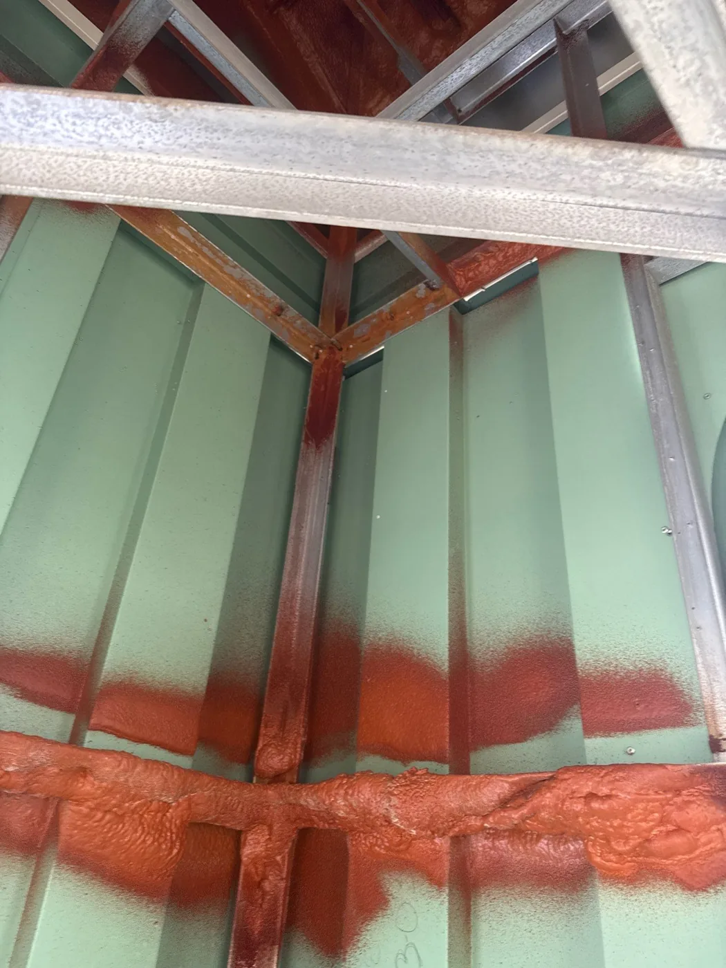 Pale green metal panels inside a steeple with bright orange spray paint marking corroded seams and a rusted joint where framing members meet.
