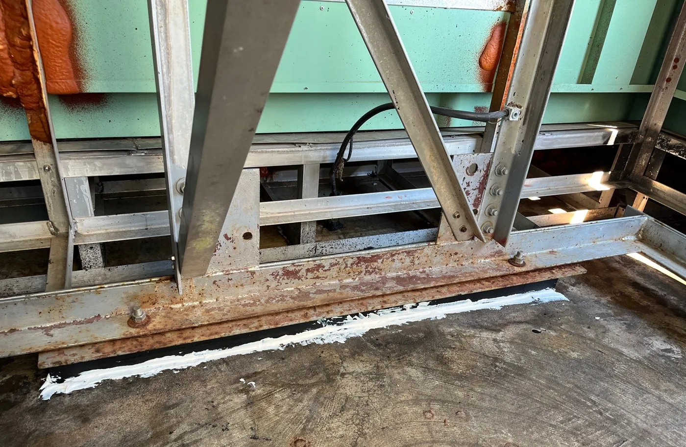 A fresh bead of white sealant applied along the joint where the rusted perimeter steel meets the deck at the base of a steeple.