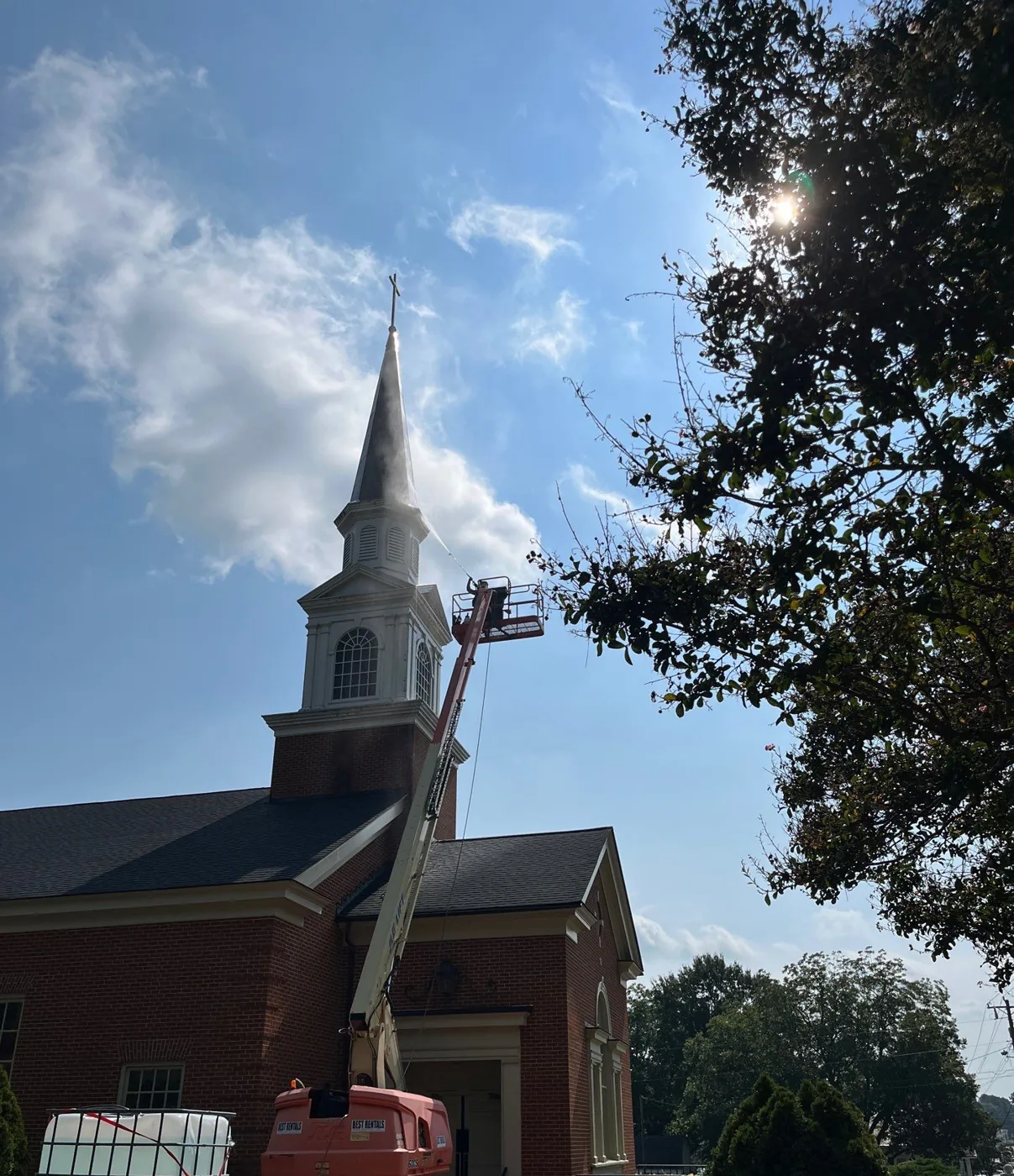 A towable boom lift parked beside a brick church with its boom raised toward the steeple, and a large water tote staged nearby.