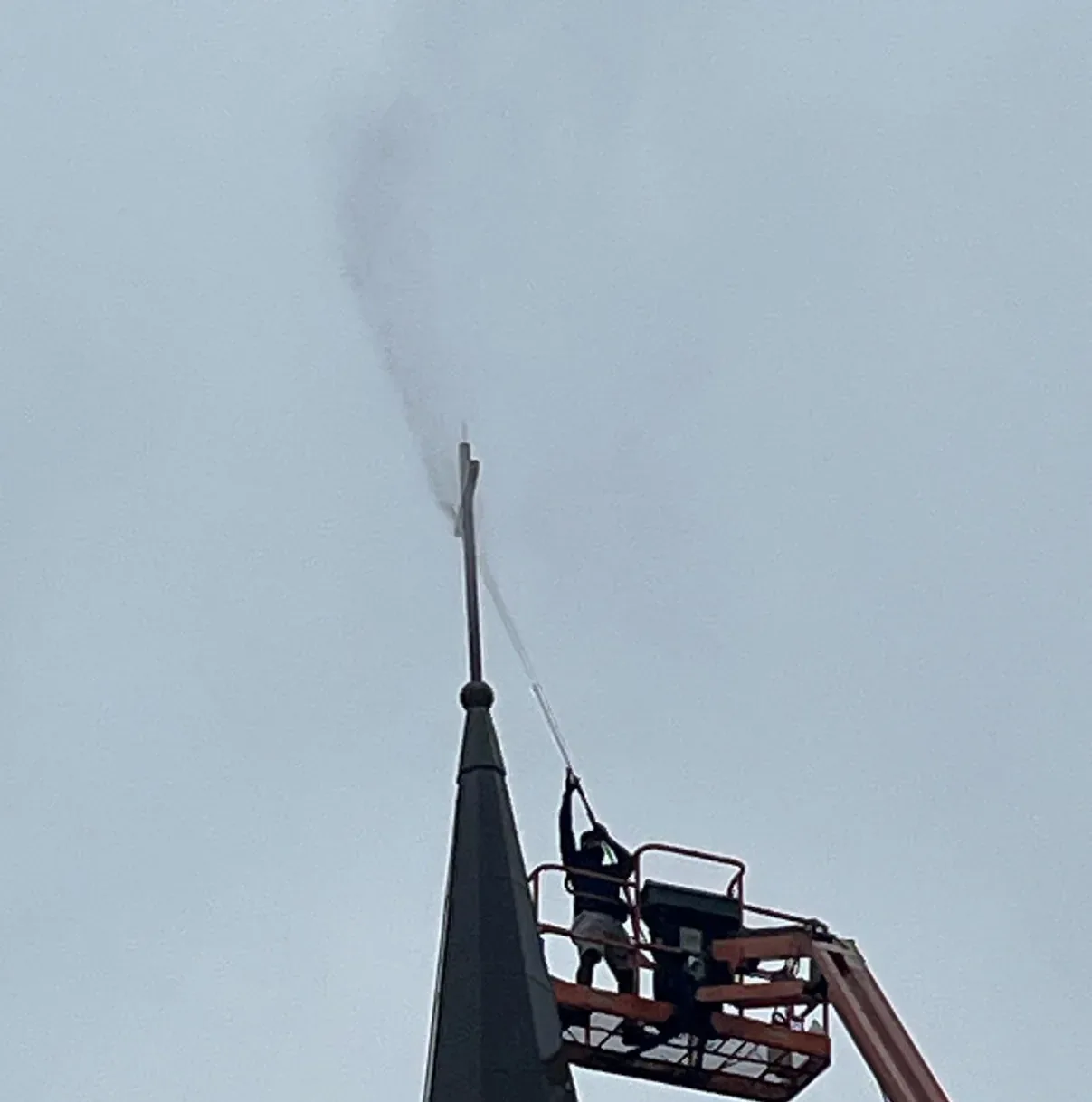 A worker standing in a raised boom lift basket beside the dark upper spire of a steeple, directing a pressure washer wand at the finial at its very tip.