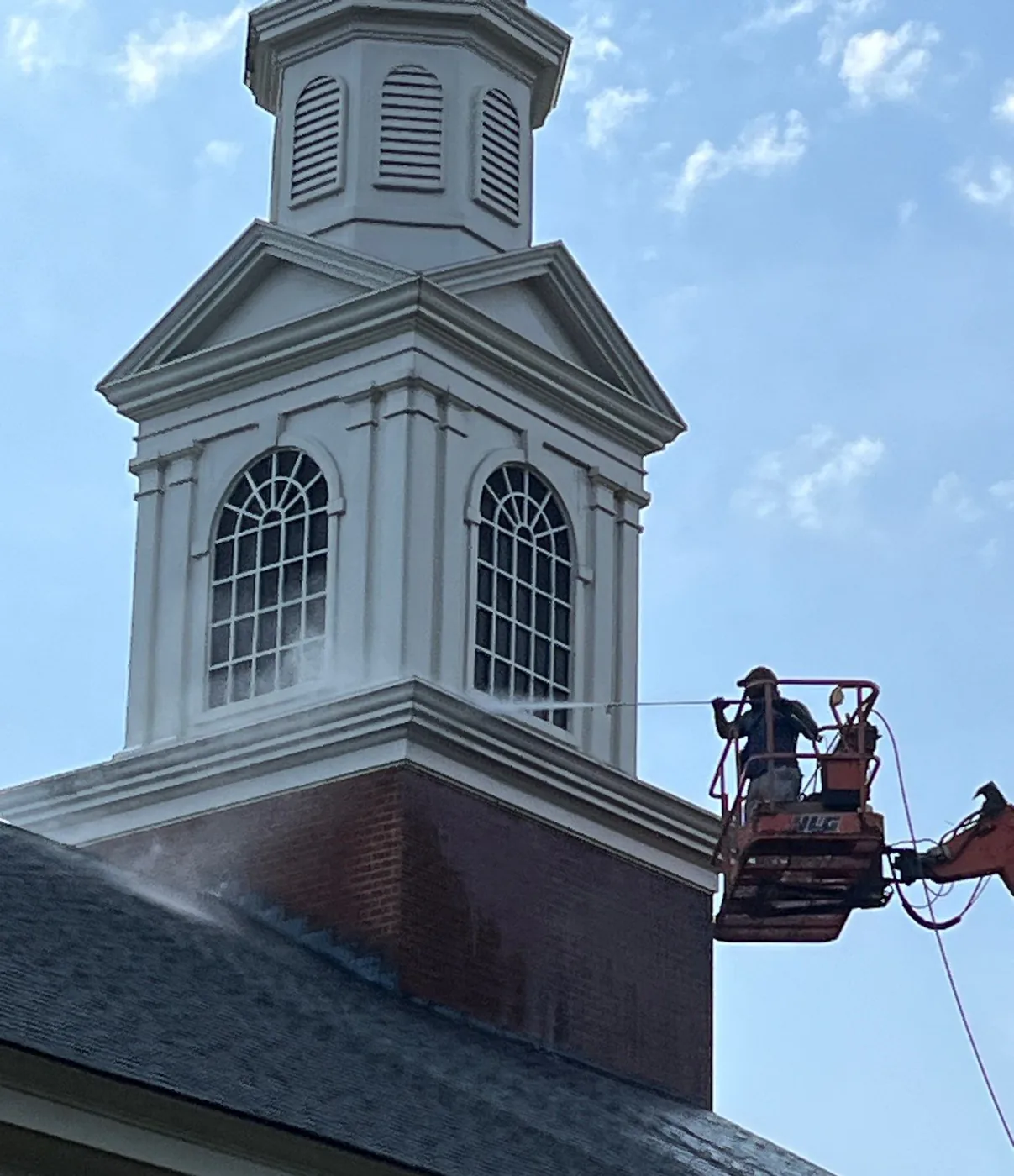 A worker in a boom lift basket pressure-washing the white louvered cupola of a church steeple, water spraying against the cornice under a blue sky.