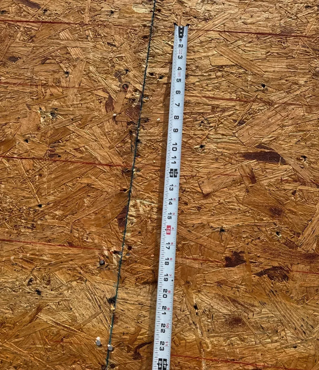 Close view of roof sheathing with a tape measure laid alongside a panel seam and a line of newly driven nails.