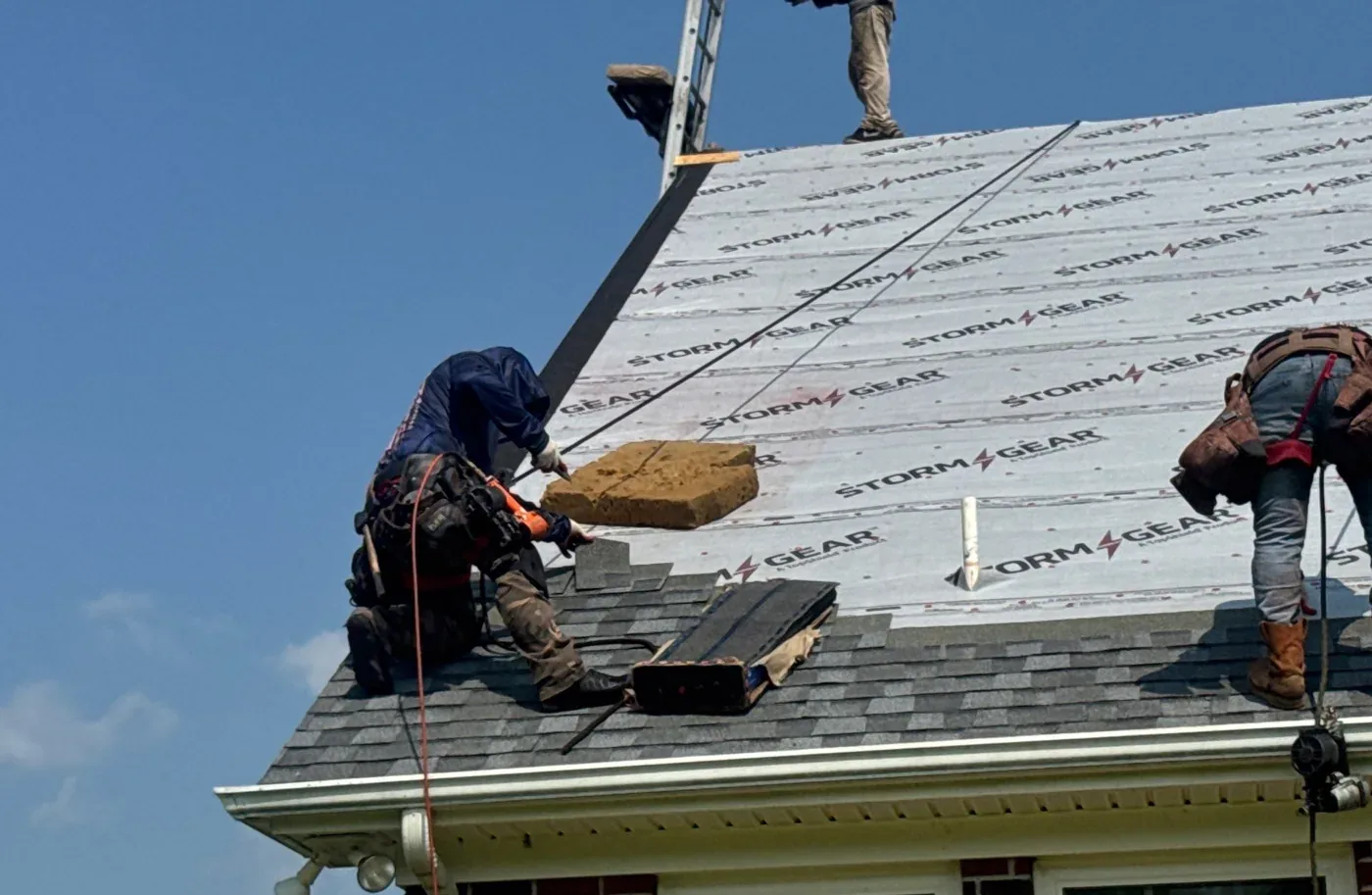 Two roofers nailing architectural shingles over synthetic underlayment at the eave of a dormer slope.