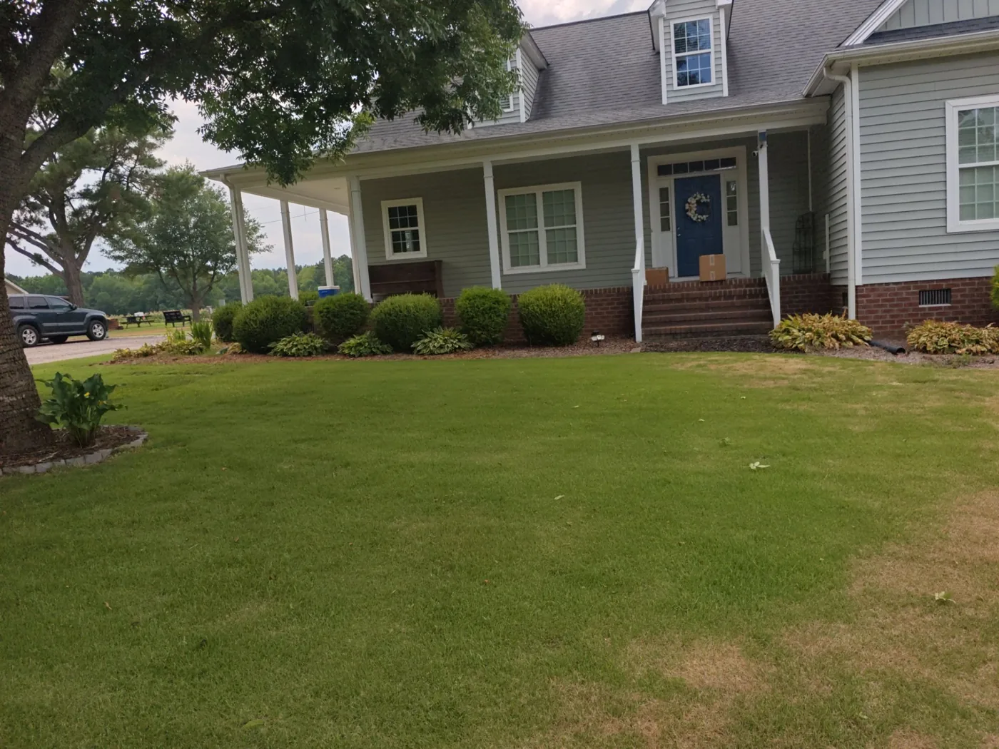 Single-story home with light green siding, a brick foundation, dormer windows and a covered front porch, photographed from the lawn before roof work began, with its original grey shingles still in place.