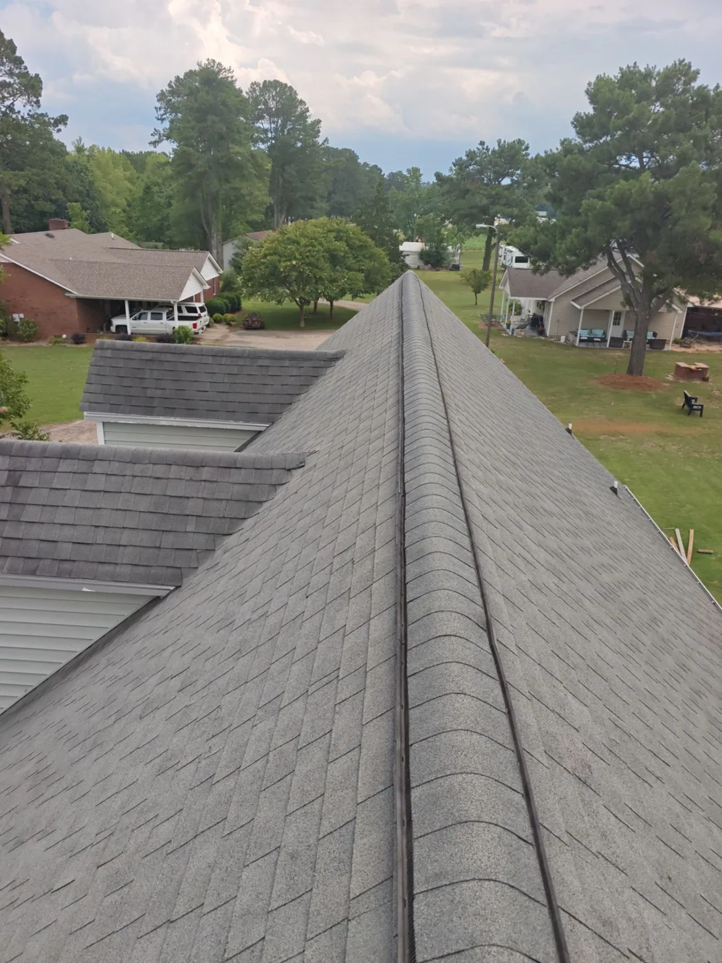 View along the full length of the original roof ridge, showing weathered grey three-tab shingles running down both slopes toward a lower dormer and the surrounding rural yard.