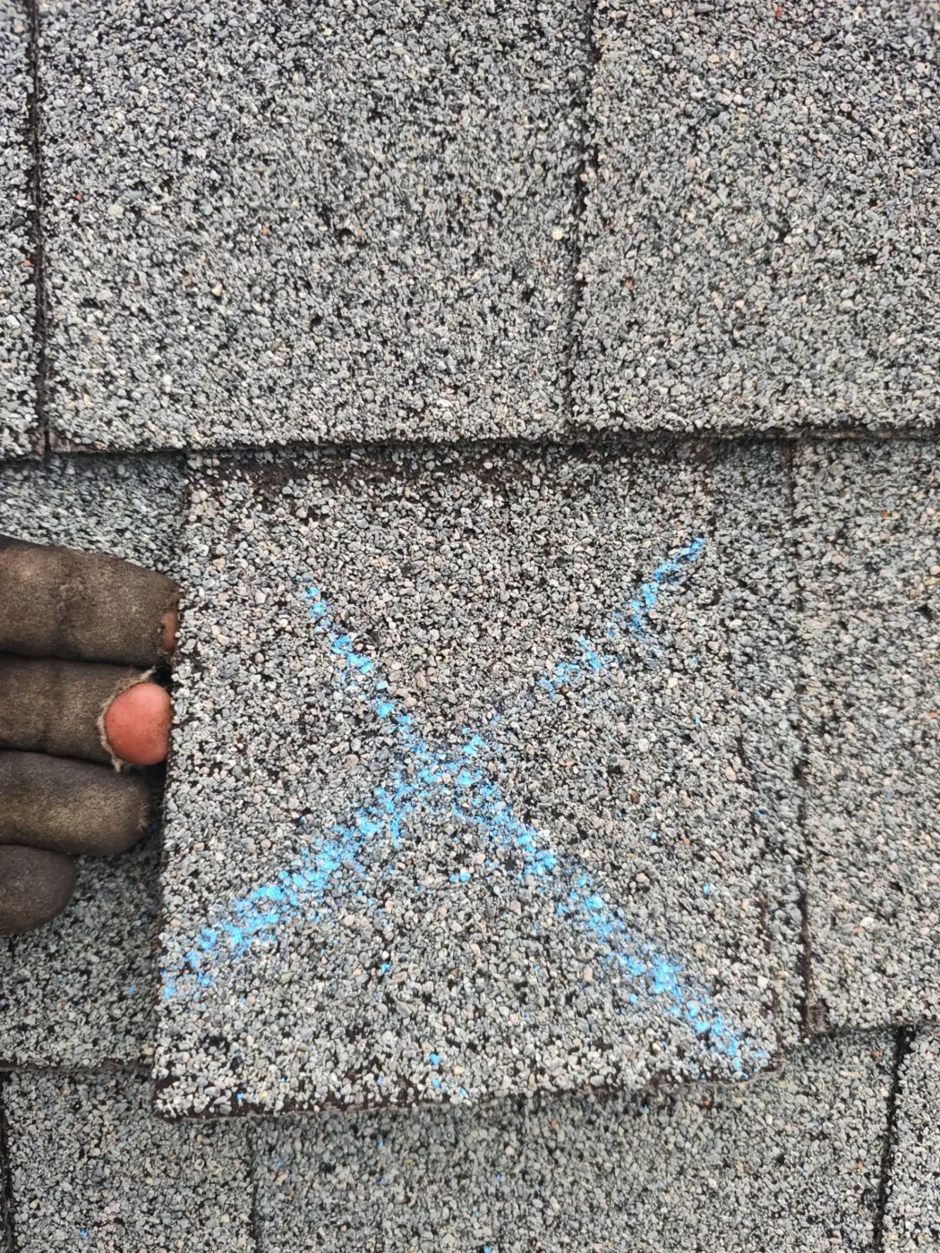 Close-up of a displaced shingle tab marked with a large blue chalk X, with a hand resting at its edge to give a sense of the size of the damaged area.