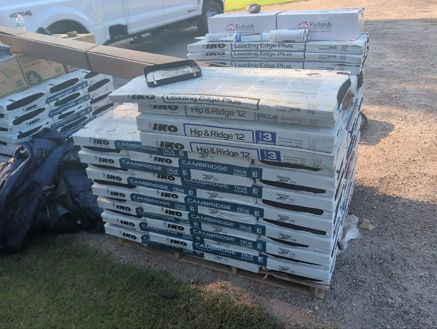 Pallet of new roofing material staged on the driveway, with wrapped bundles of IKO Cambridge architectural shingles in charcoal grey, IKO Hip and Ridge 12 caps and Leading Edge Plus starter strip.