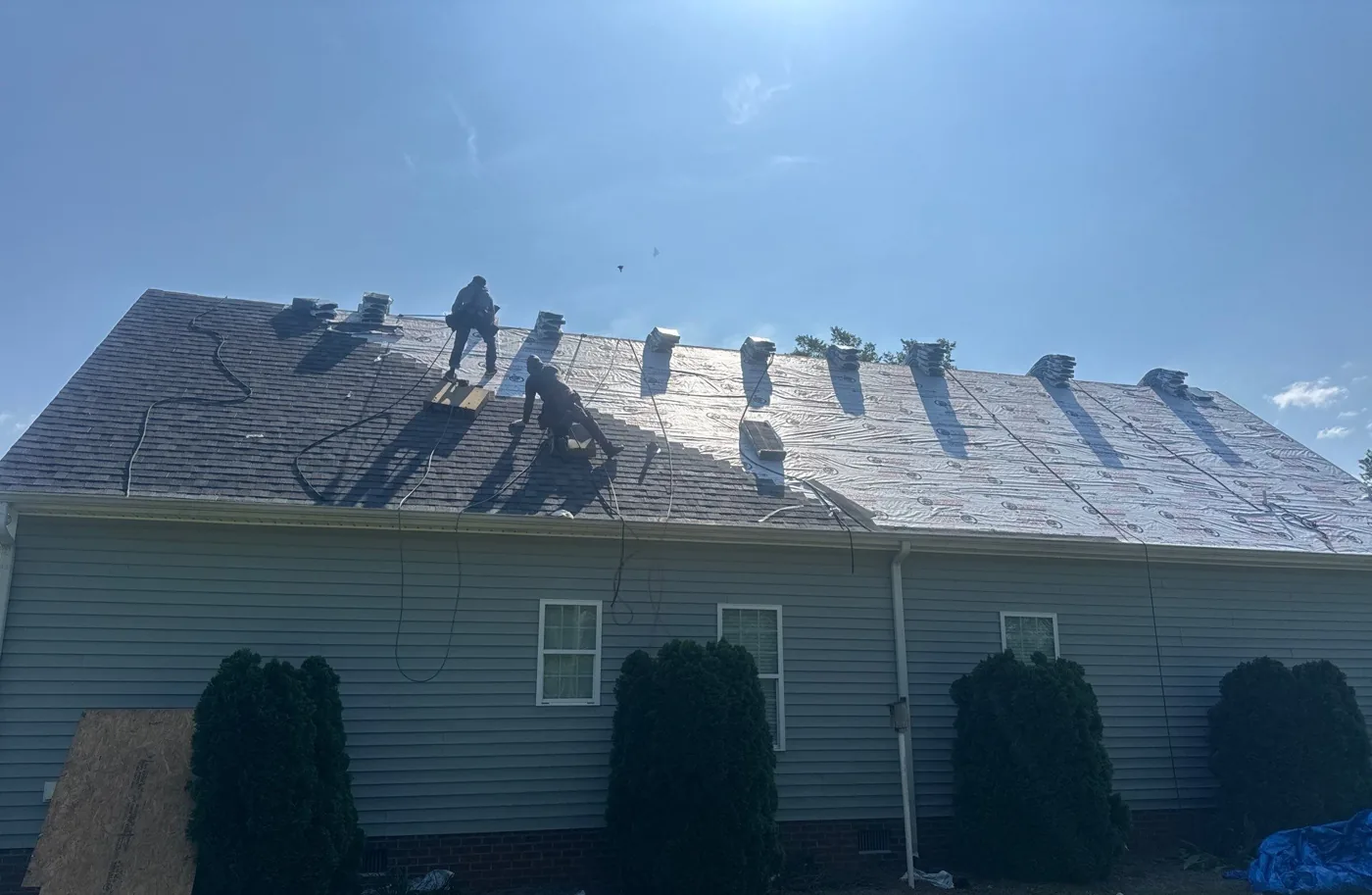 Rear slope of the house mid tear-off, with two crew members working the line between stripped and unstripped roof, old shingles cleared to the left, synthetic underlayment rolled out to the right and new bundles staged across the deck.