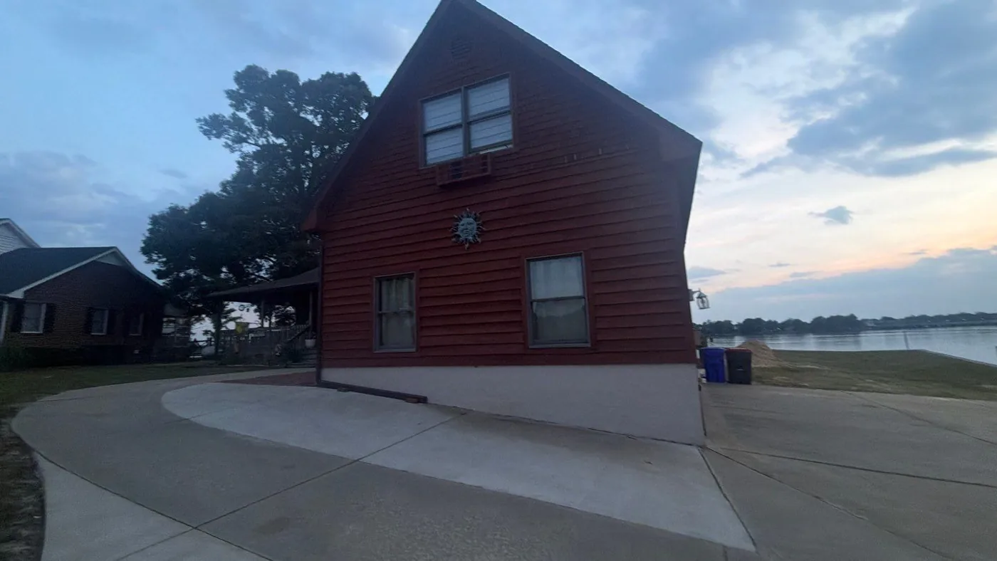 Gable end of a red-stained wood-sided waterfront home at dusk, still covered by its original dark, aged shingle roof, with open water visible beyond the lawn.
