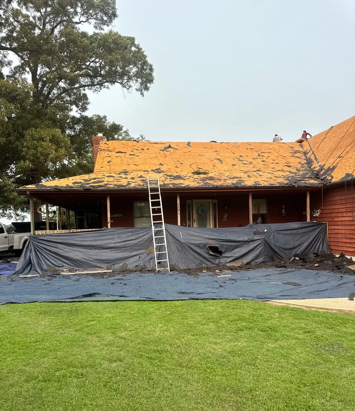 The long low roof of the home part-way through tear-off, bare plywood showing across the stripped section, with dark tarps hung down the walls and over the landscaping below.