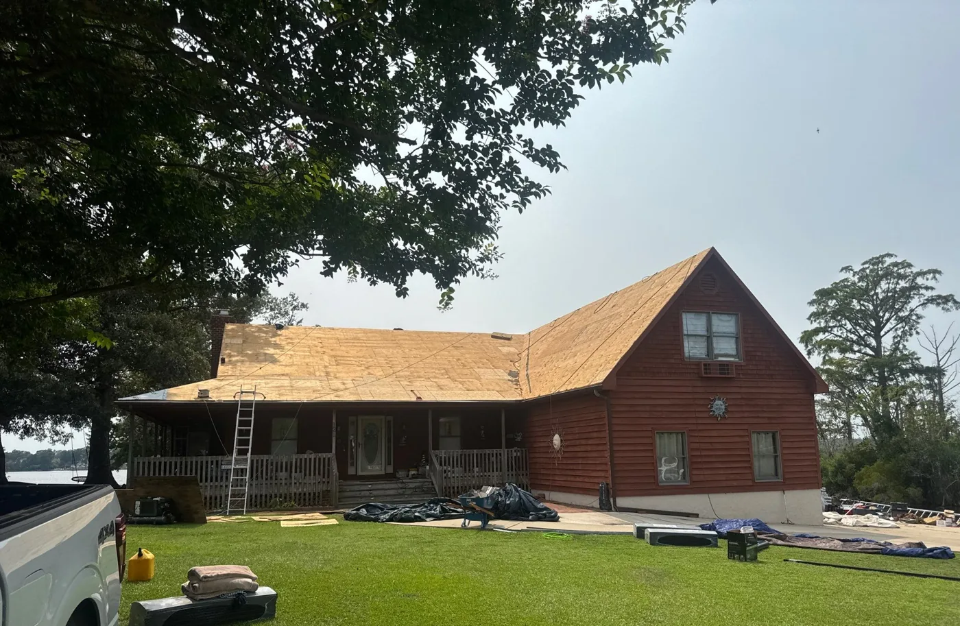 Gable end of the home with the roof stripped completely to bare plywood sheathing, a ladder at the eave and new roofing materials staged on the driveway below.