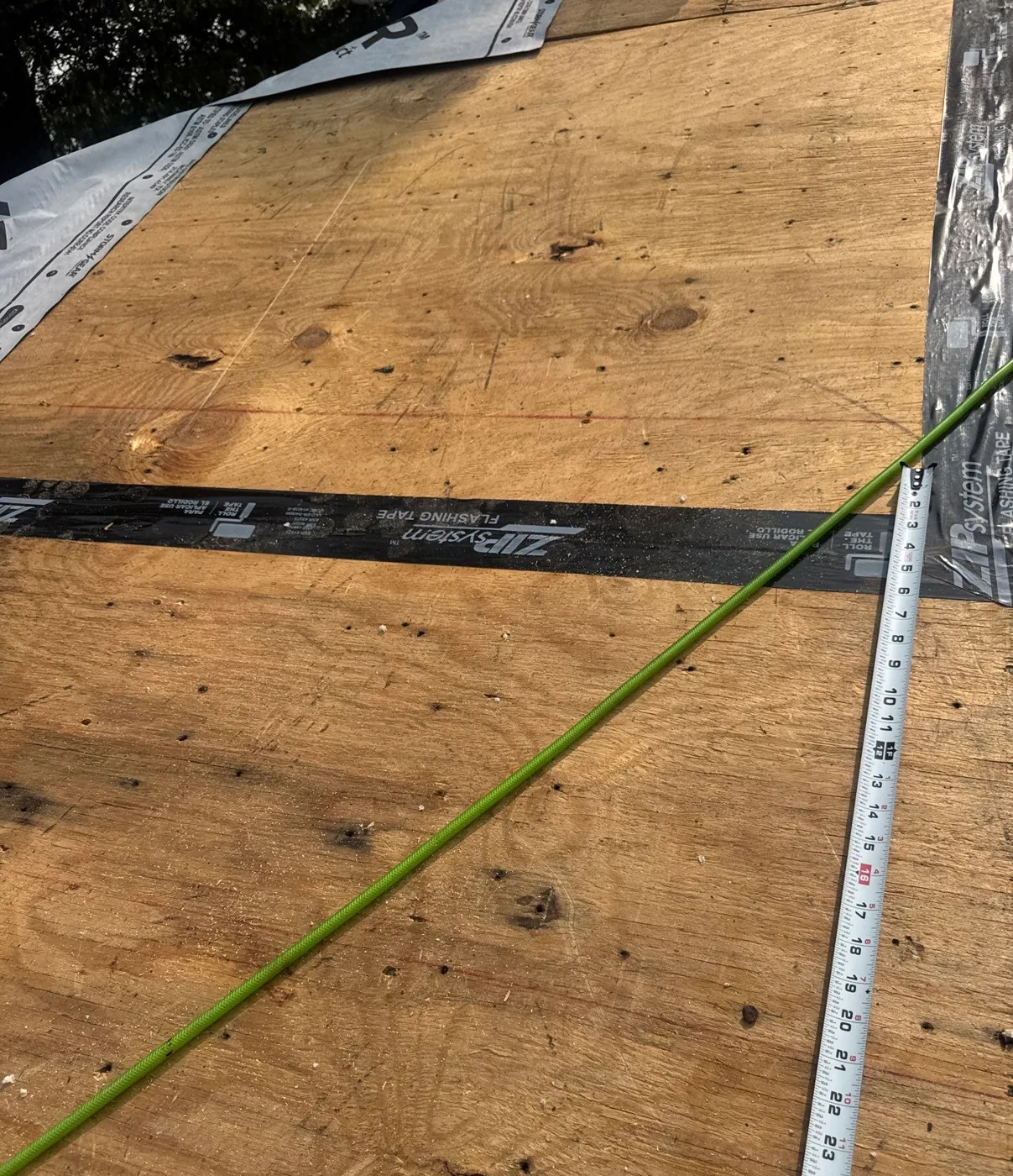 Close-up of the plywood roof deck with a black ZIP System flashing tape strip pressed over the seam between sheets, chalk lines snapped across the deck and synthetic underlayment being rolled over the top.