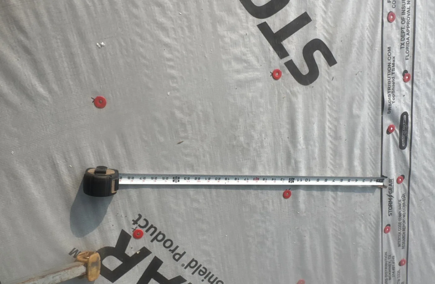 Synthetic underlayment fastened over the roof deck with red plastic cap nails, a steel tape measure extended across the sheet to check the spacing between fasteners.