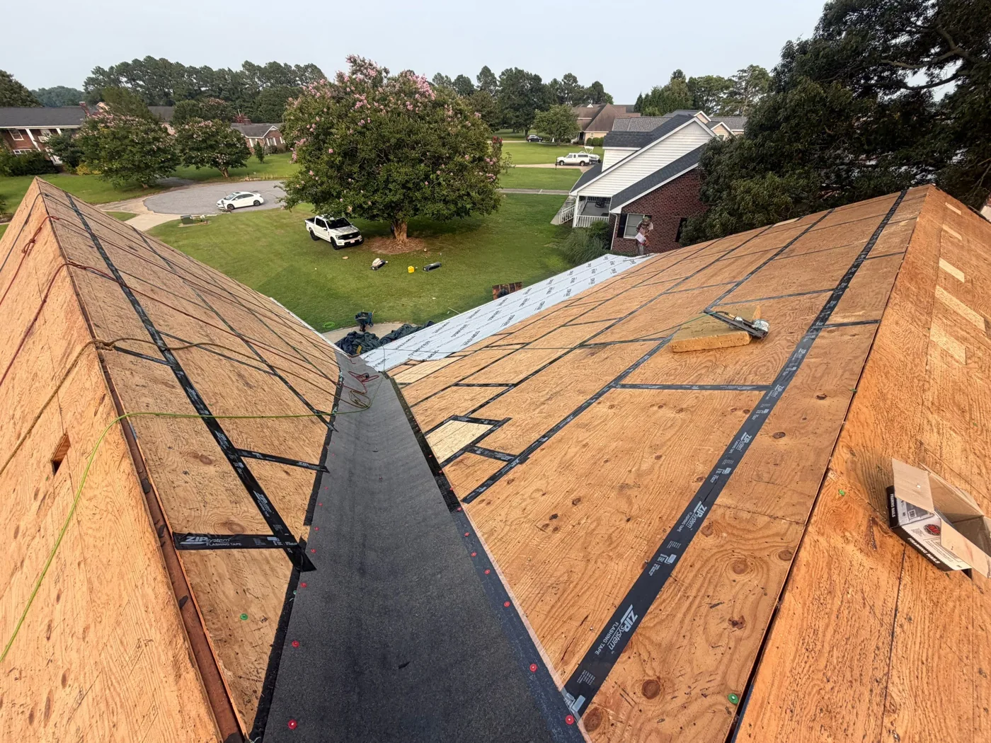 View down a roof valley between two slopes of bare plywood decking, with a black self-adhering membrane run the full length of the valley and black seam tape applied along the deck joints on both sides.