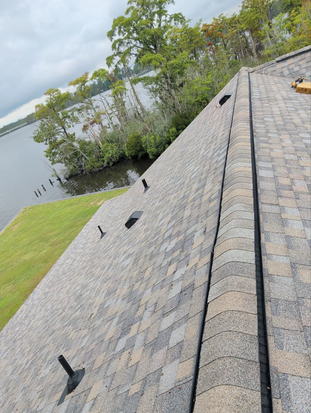 Finished grey architectural shingles running up to a capped ridge, with two low-profile black roof vents set into the slope and open water visible beyond the lawn.