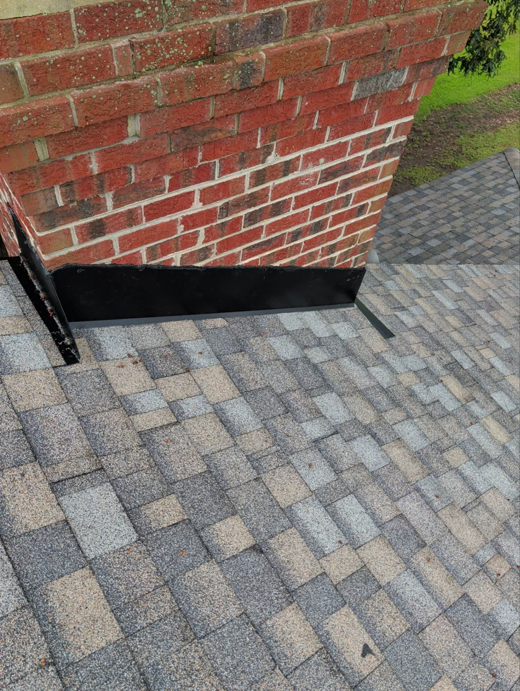 Close view of the base of a red brick chimney where it meets the new shingle roof, with dark metal flashing fitted tight along the joint.