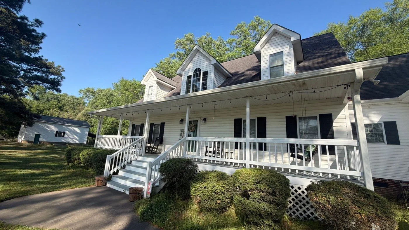 Two-story white-sided home with black shutters, dormers and a full-width covered front porch, photographed before roof work with its original dark, weathered shingles in place.