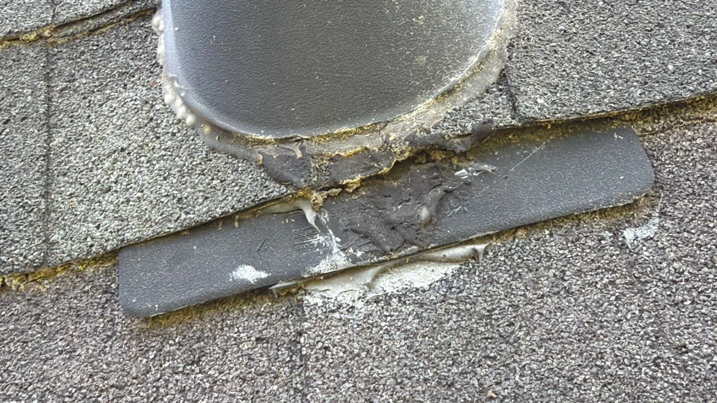 Close-up of an aged plumbing vent pipe on the old roof, with a split, chalky bead of sealant and a cracked shingle at the base of the boot.