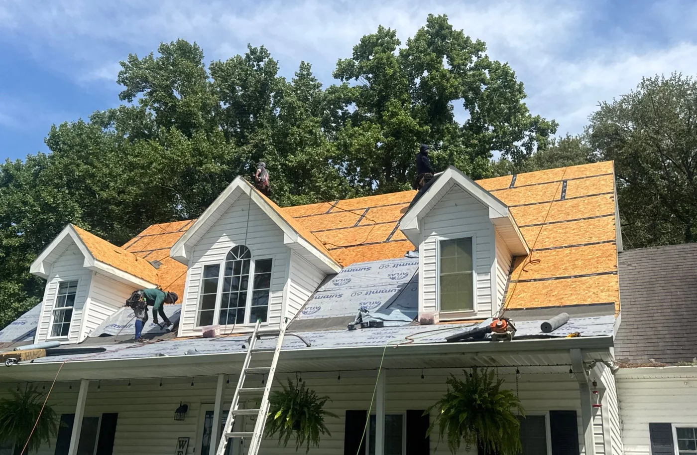 The main roof stripped down to bare orange OSB decking across all slopes and dormers, with a ladder at the eave and crew members working on the exposed deck.