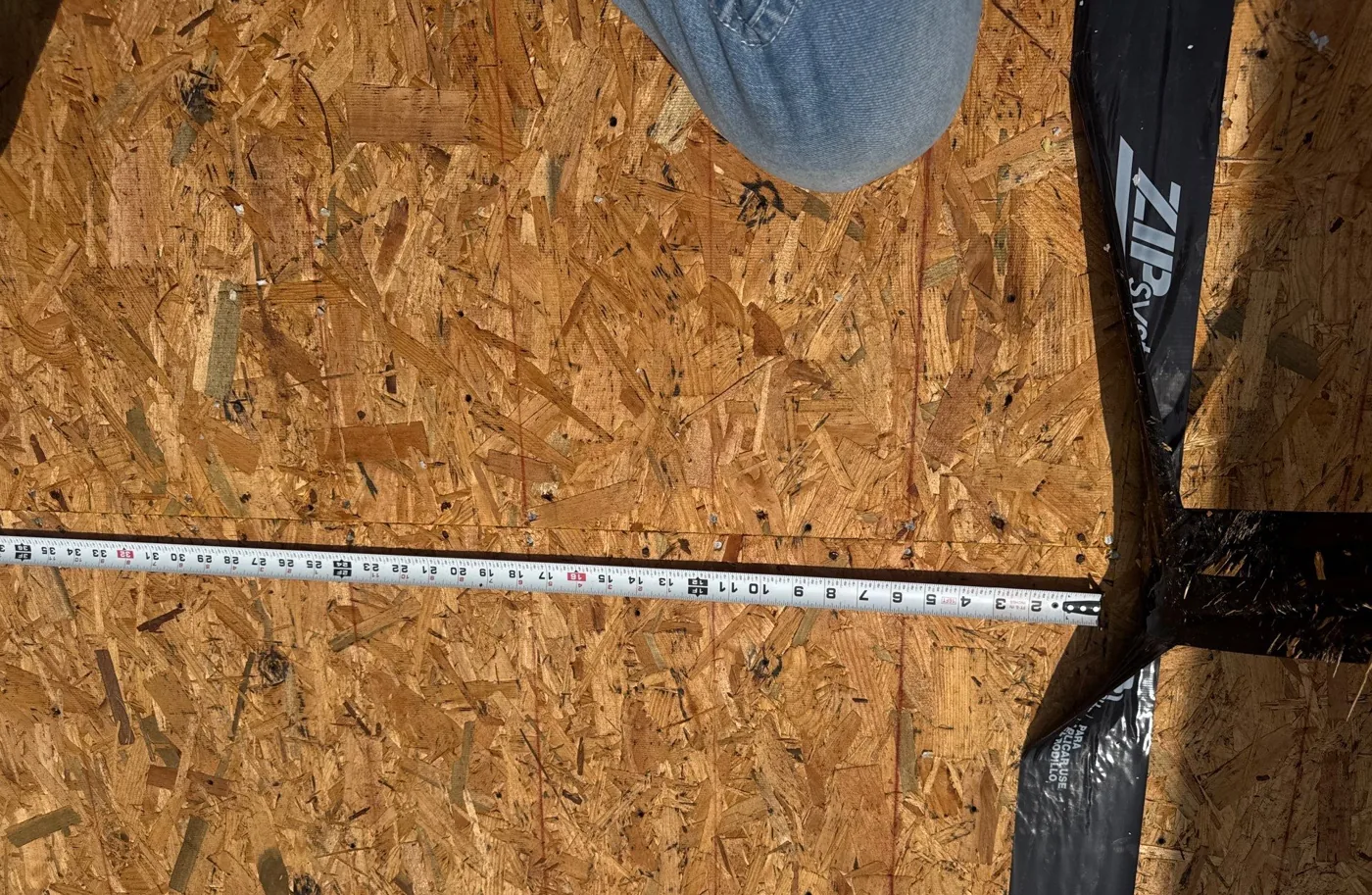 Close-up of exposed OSB roof decking with black ZIP System seam sealing tape applied over the joints between sheets, a tape measure laid across the deck for scale.