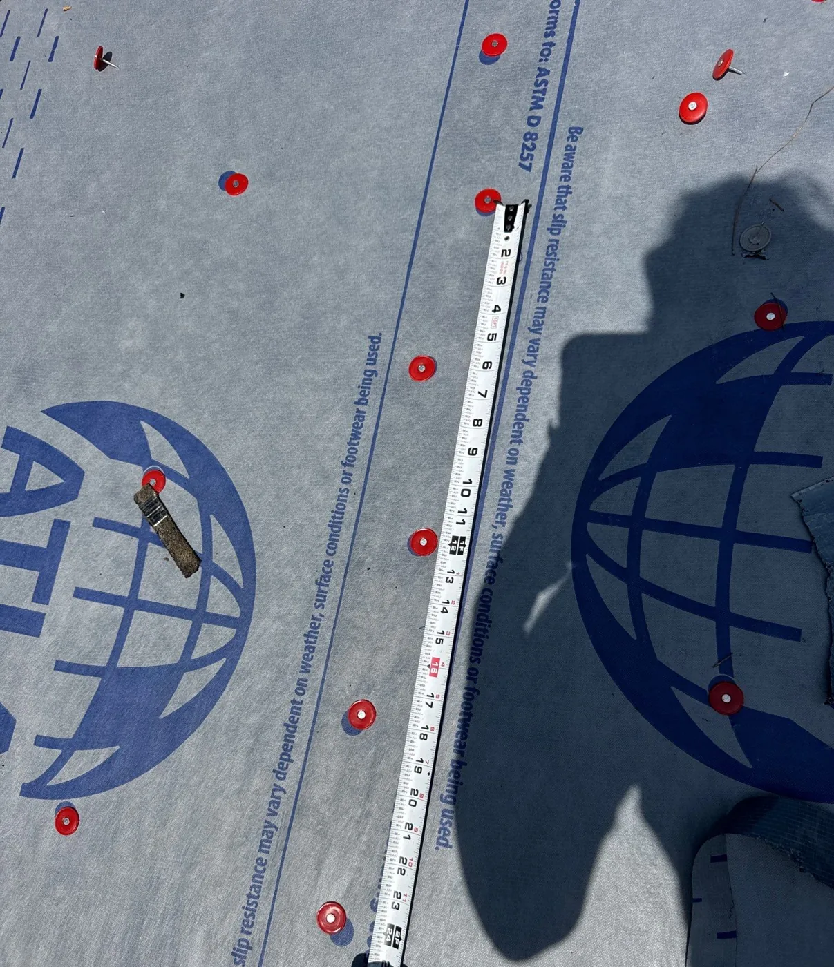 Synthetic underlayment rolled out over the roof deck and fastened with evenly spaced orange plastic cap nails, with a tape measure laid across it to show the fastener spacing.