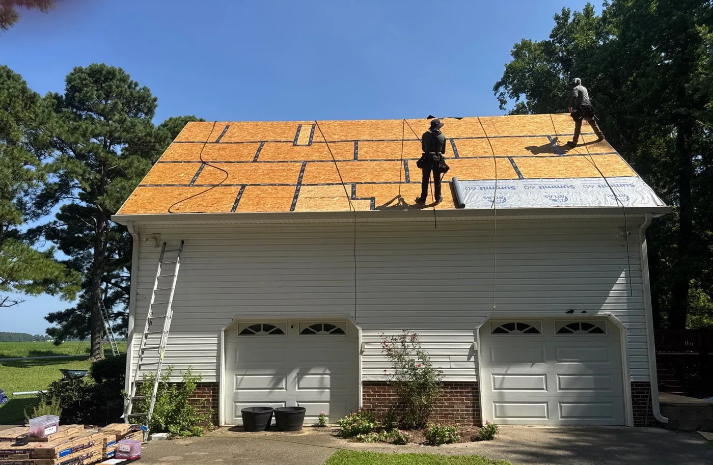 Detached three-bay garage with its roof stripped to bare OSB decking, a crew member standing on the exposed deck and a ladder set against the eave.