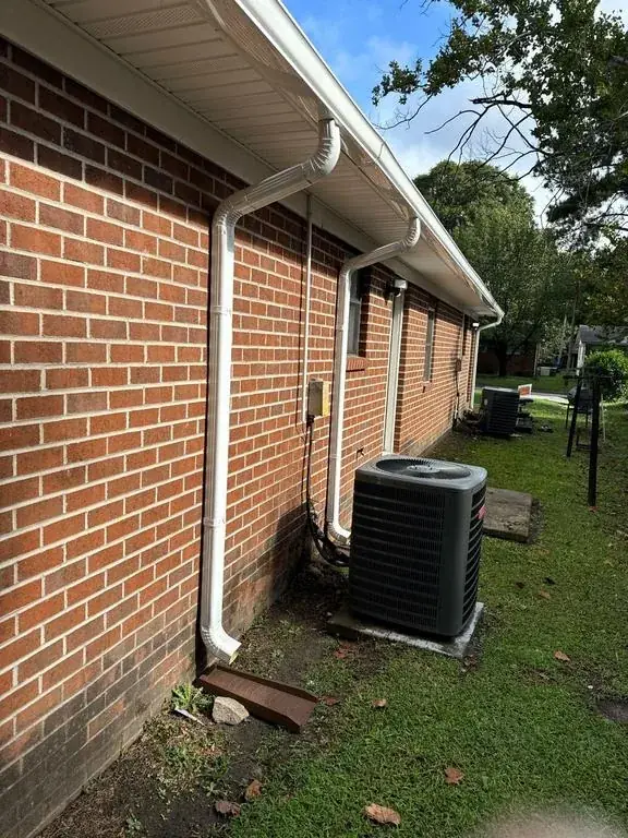 White downspouts on a brick wall, one routed to land beside an air-conditioning unit rather than onto it