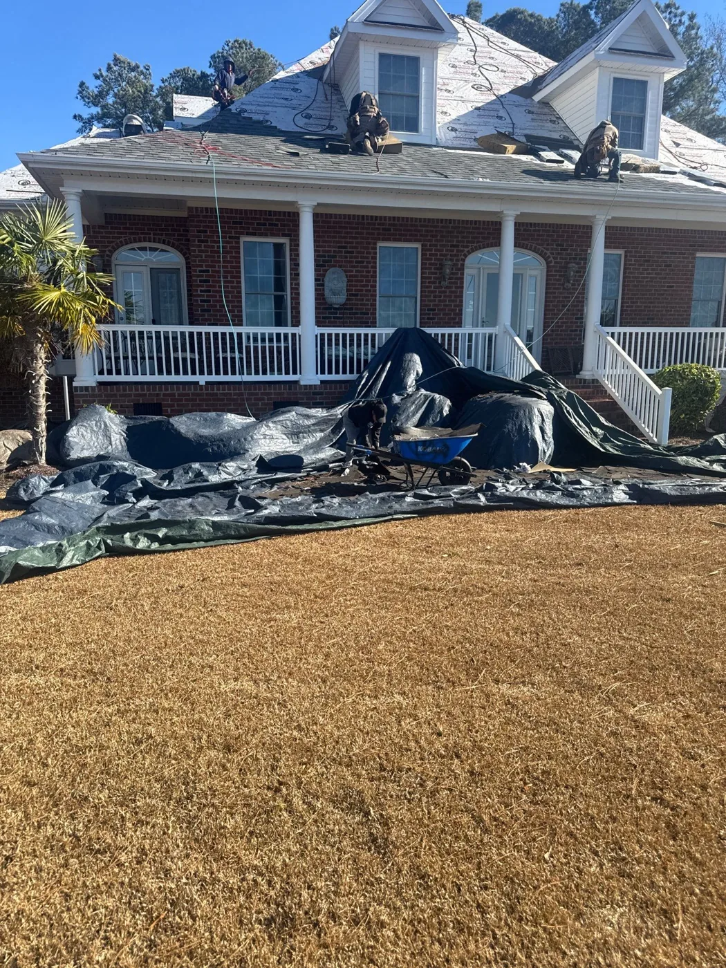 A brick colonial home mid re-roof, the old shingles stripped to the deck with the crew working on the roof and tarps protecting the lawn and shrubs below