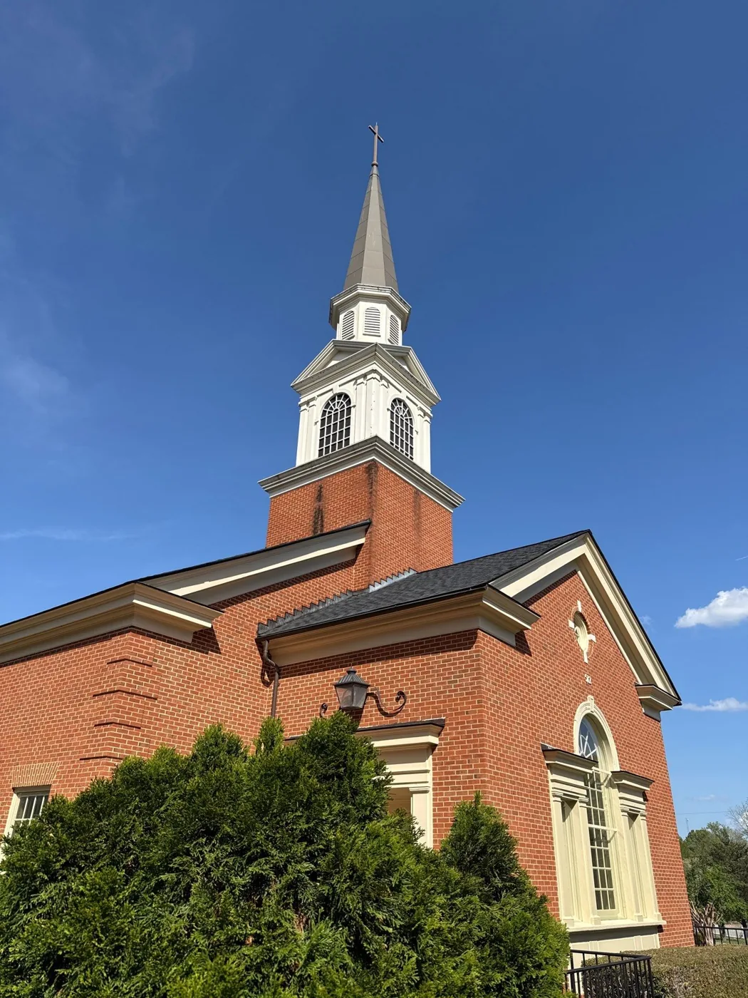 A brick church with a white belfry, louvered lantern and tall grey spire topped by a cross against a deep blue sky