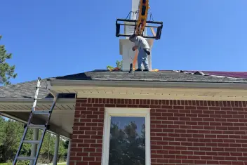 Telehandler holding a church steeple up while a crew member works on its base on a brick church roof